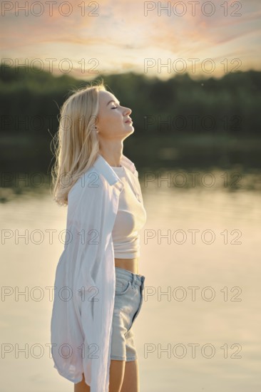 A woman stands by the calm lake, feeling the gentle breeze as she admires the colorful sunset sky.