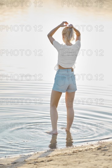 A woman stands in shallow water, playfully wringing her hair as sunlight glimmers on the surface.