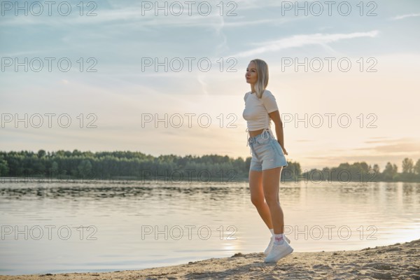 A young woman walks leisurely along the sandy lakeshore as the sun sets behind the trees. She wears