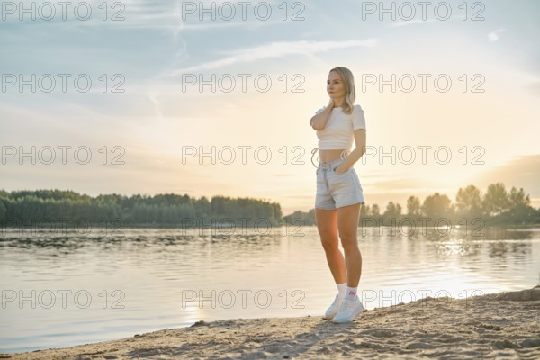 A young woman relaxing on the sandy beach near a calm river at sunset. She wears a casual outfit