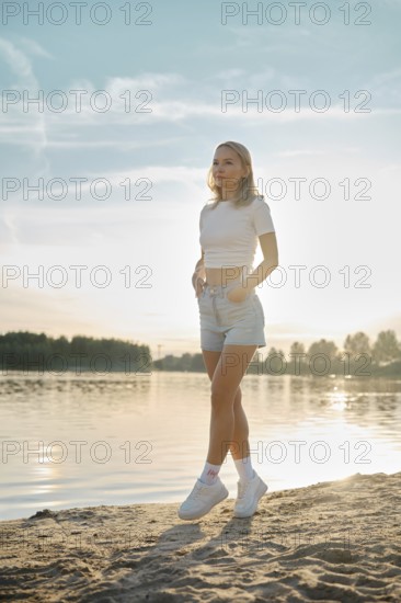 A young woman walks on tiptoes on a sandy shore as the sun sets over the lake. Her casual outfit