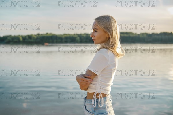 A blonde woman stands in profile by a calm lake, gazing thoughtfully into the distance. The sun