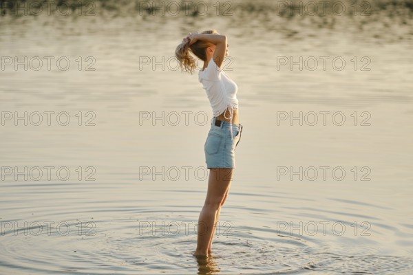 A woman stands in shallow water in profile, basking in the warm glow of sunset. She raises her