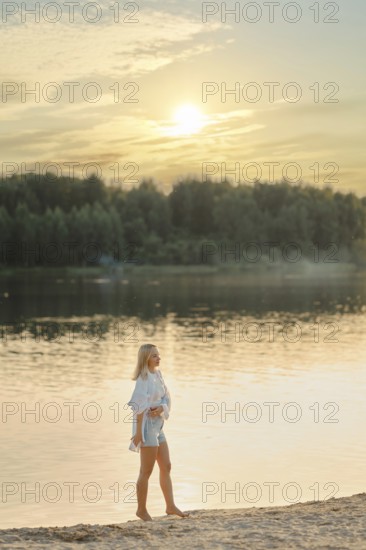 A woman strolls barefoot along the sandy shore of a serene lake during sunset. The sun sets behind