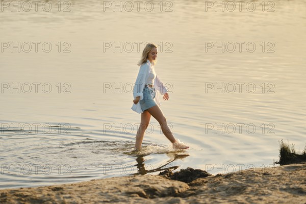 A young woman with blonde hair strolls through shallow water during sunset, her feet gently making