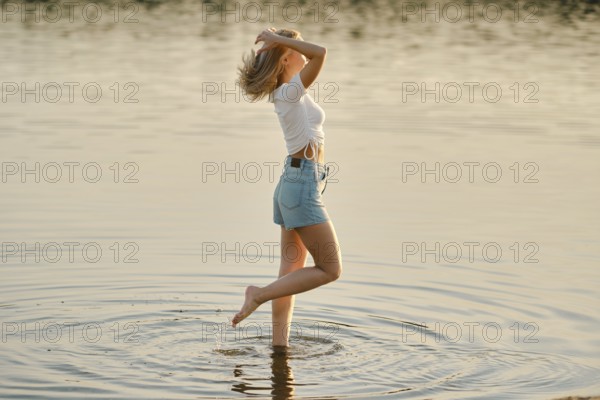 A young woman is joyfully splashing in the shallow waters of a serene lake. The sun is setting,