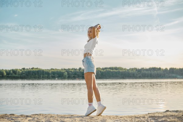 A young woman walking along a tranquil lake at sunset, dressed in casual summer attire. She