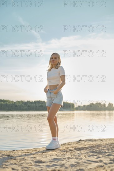 A young woman stands on a sandy beach, facing the camera. She wears a casual outfit of a cropped