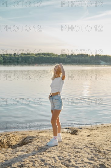 A young woman stands on the sandy shore of a calm lake, admiring the sunset. Her relaxed pose and