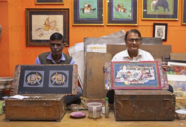 Miniature painters, aged 50 and 52, show paintings of elephant and royal city palace in studio,