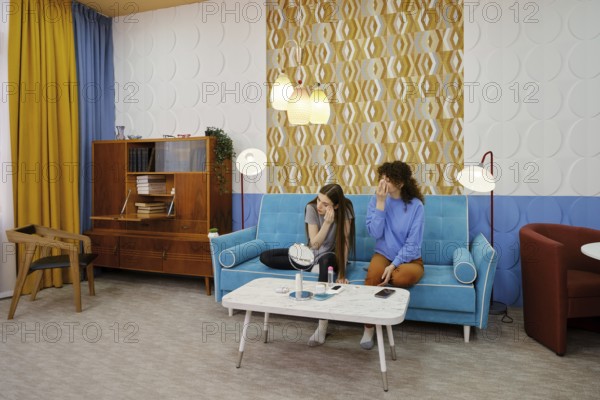 Two women sit on a turquoise sofa in a stylish living room and removing makeup. The vibrant decor