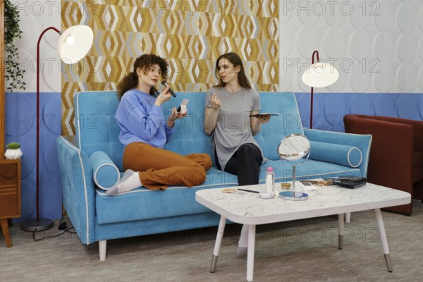 Two friends are sitting on a blue sofa and discussing makeup. The room features stylish decor,