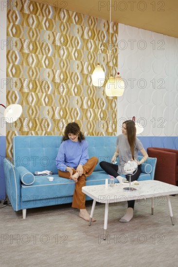 Two young women are engaged in skin care procedures at home sitting on a cozy sofa. One woman rubs