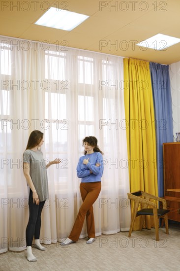 Two women are talking in a bright room featuring large windows with flowing curtains in yellow and