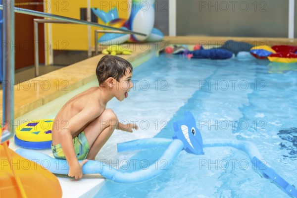 Happy child crouching at the edge of an indoor swimming pool, playing with a pool noodle shaped