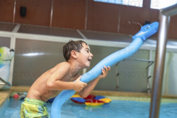 Happy child playing with a pool noodle shaped like a whale and spraying water in an indoor swimming