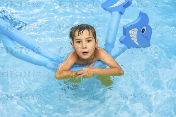Young boy playing with a shark shaped pool noodle float in a blue swimming pool, enjoying his