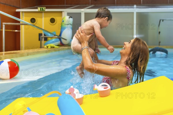 Smiling mother lifting her baby while playing in a swimming pool during a sunny day, enjoying their