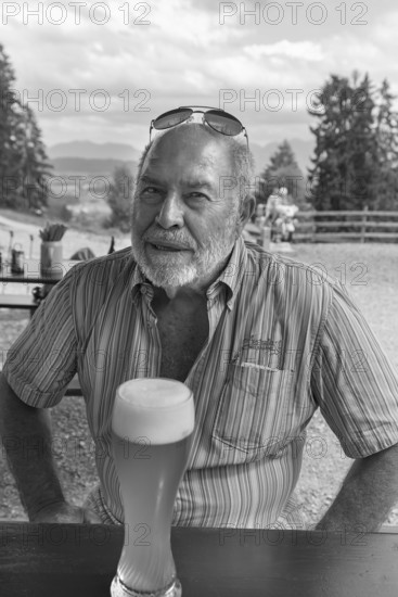 Elderly gentleman with beard and sunglasses in a garden restaurant with a drink, black and white,