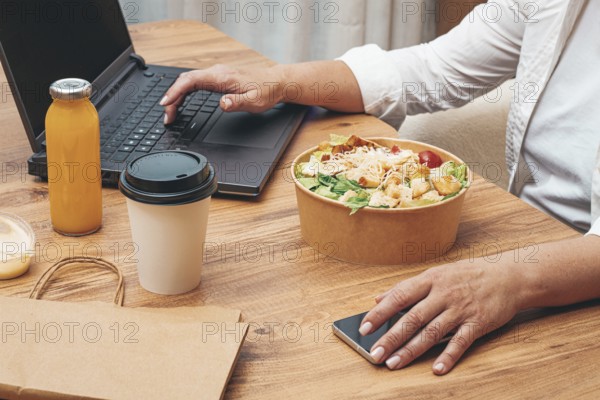 A woman is working on a laptop at home, there is food on the table, salad, coffee and juice,