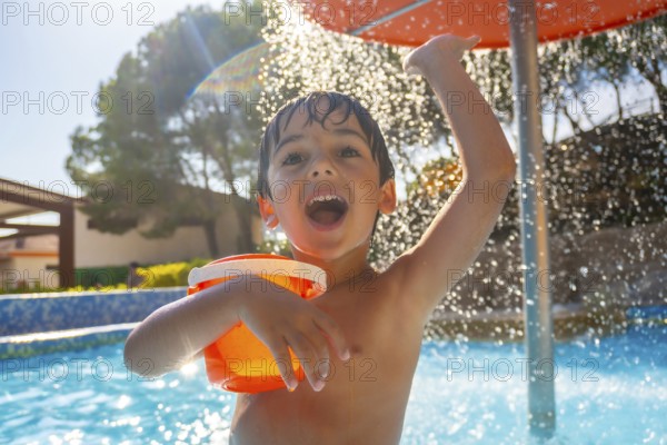 Boy holding orange bucket and raising arm under refreshing water shower in swimming pool on sunny