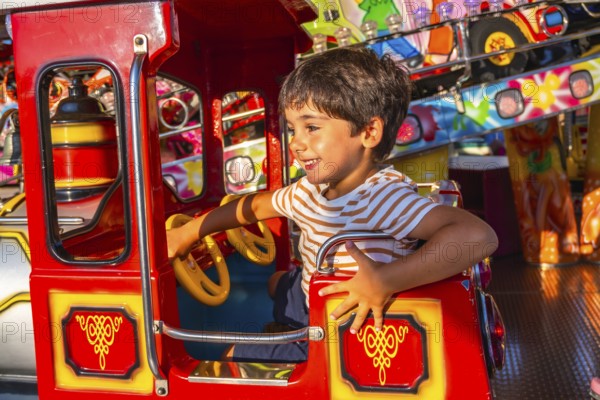 Young boy smiling while steering a vibrant red amusement park ride, surrounded by colorful