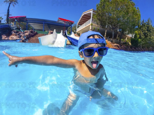 Playful child with blue sunglasses and hat pointing with finger and sticking tongue out in a