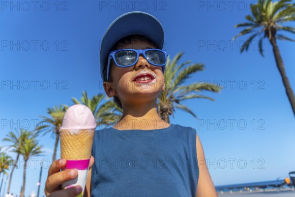 Young boy wearing sunglasses and a cap enjoys a strawberry ice cream cone on a sunny beach with