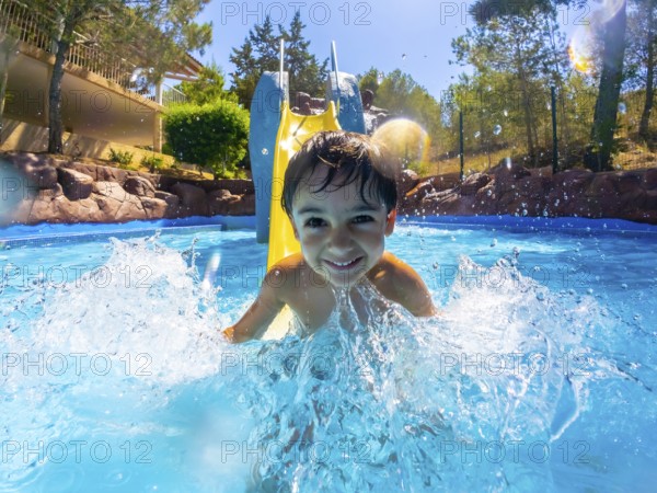 Young boy enjoying a sunny day, sliding into a vibrant blue pool with a big splash, surrounded by
