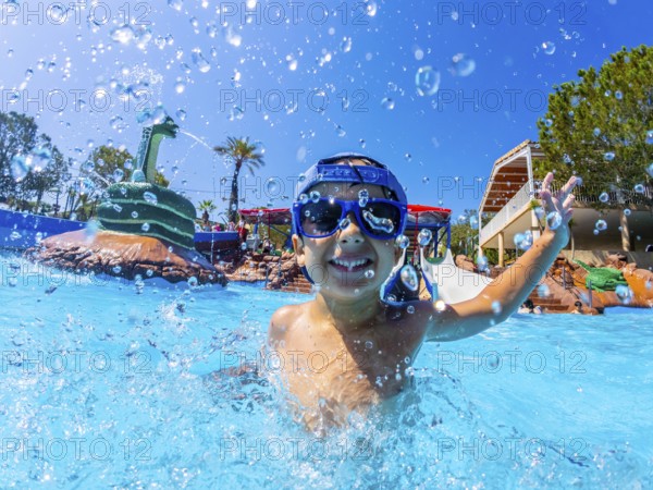 Happy boy wearing swimming goggles, splashing joyfully in a vibrant water park pool during summer