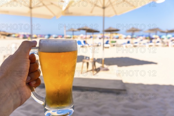 Tourist enjoying refreshing beer on sunny beach with blurred umbrellas and ocean, perfect summer