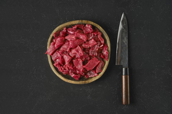 A wooden bowl holds freshly cut pieces of raw beef, showcasing a vibrant red color. Beside the