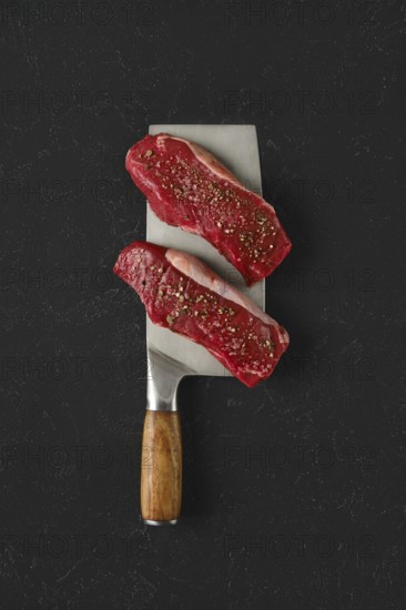 Two raw beef steaks seasoned with spices on a large metal cleaver with a wooden handle. The dark