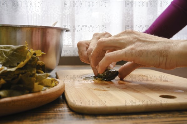 Hands delicately wrap seasoned filling in grape leaves on a wooden cutting board. Fresh ingredients