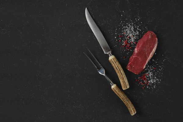 A knife and fork, with horn handles, rest beside a raw beef steak on a dark surface. Salt and pink