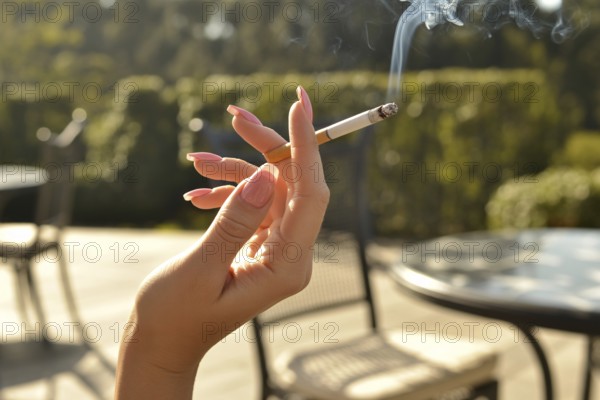 Elegant female hand with long pink nails holding a burning cigarette. Summer sunlight in outdoor
