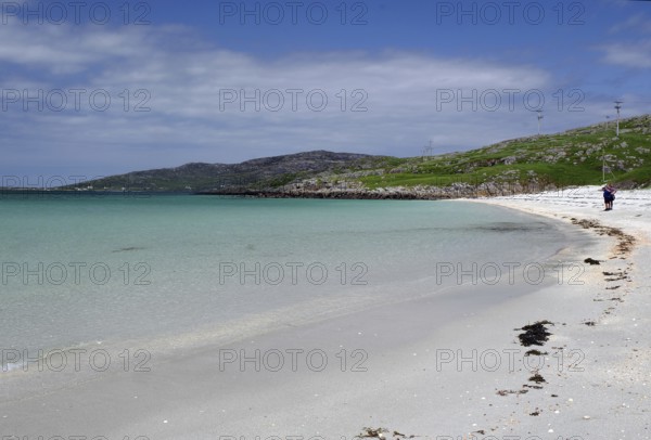 Clear bright blue beach with gentle waves under a cloudless sky, two people at the water's edge,