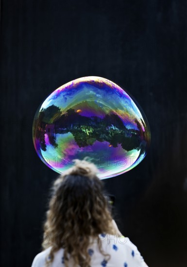 A woman looks at a large iridescent and floating soap bubble in the colours of the rainbow against