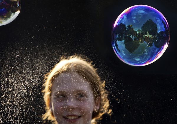 A girl is happy about a shimmering and floating soap bubble in front of a black background,