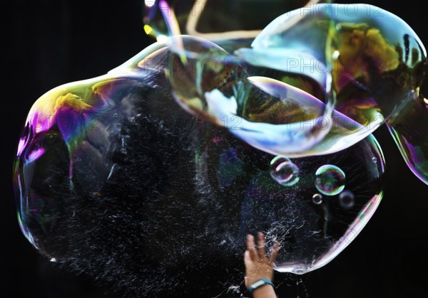 A hand bursts a large iridescent soap bubble against a black background, Recklinghausen, Germany