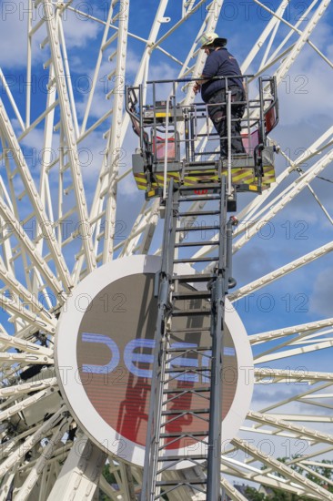 A worker stands on a ladder in front of a large Ferris wheel under a blue sky, fire brigade
