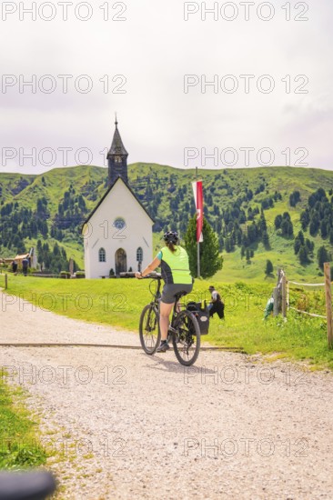 A cyclist rides along a gravel path, in the background a village church and green hills, Zallinger