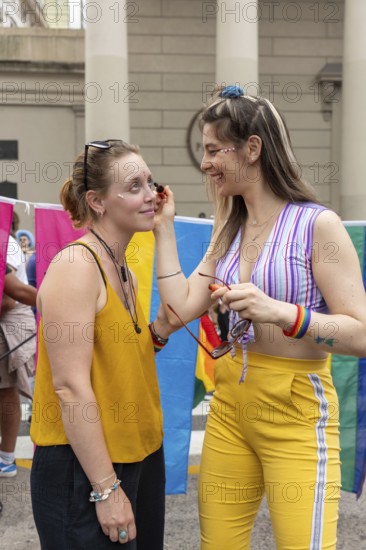 During a vibrant gay pride event, a woman applying make-up to her friend's face highlights the