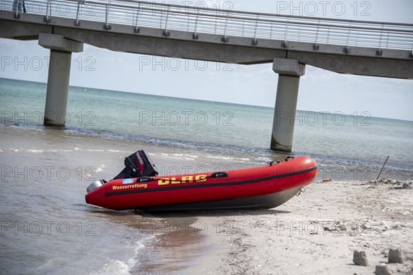 A DLRG lifeboat lies on the beach in front of a bridge with walkers and the sea in the background,
