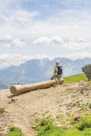 A hiker sits on a tree trunk and looks at the surrounding mountains, Schlehrn hike, Alpe di Siusi,