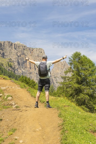 A man stands joyfully with outstretched arms on a sunny mountain path, Schlehrn hike, Alpe di