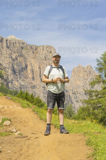 A man with a rucksack stands smiling on a mountain path with a rocky background, Schlehrn hike,