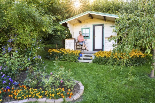 A woman sits relaxed on the small veranda of a garden shed and reads. Blooming summer flowers in