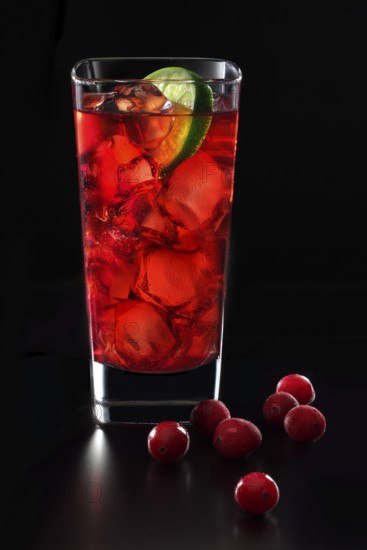 Cocktail in a long drink glass on ice with a slice of lime and cranberries as garnish against a