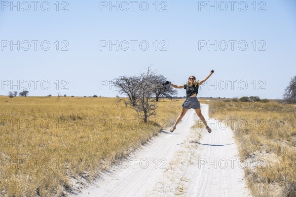 Tourist with camera jumping on a sandy track between yellow dry grass, Makgadikgadi salt pans,